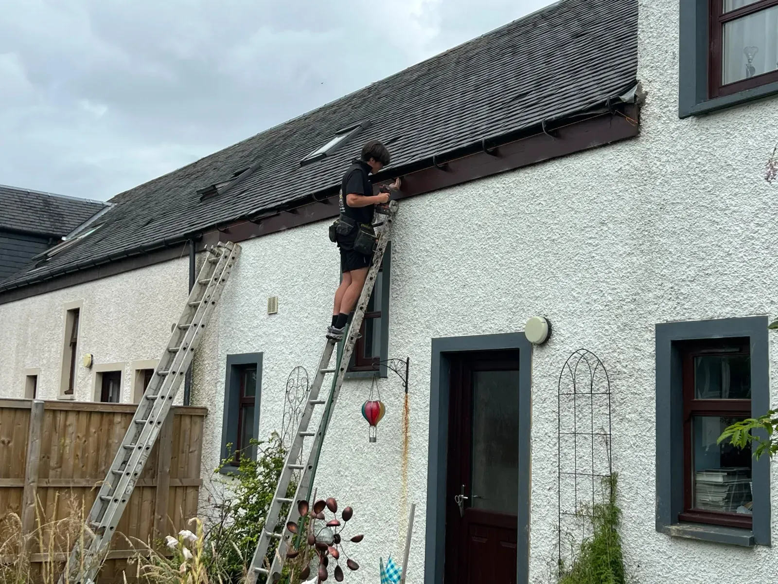 Roofer on a ladder working on the eaves and guttering of a white-rendered cottage